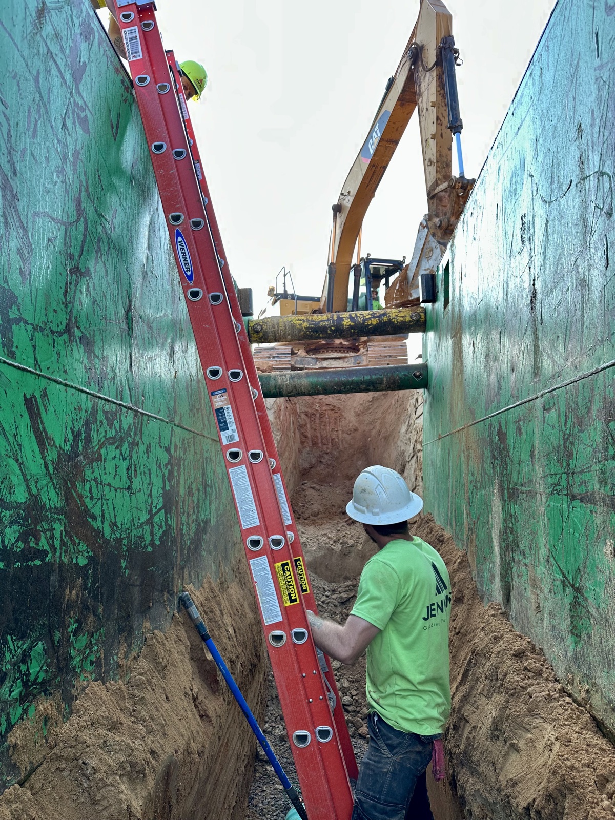 Jenkins crew member in a shored utility trench with excavator overhead